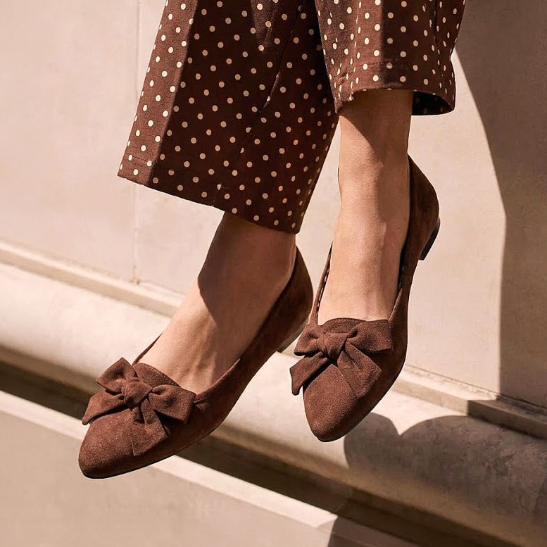 A person wearing brown suede ballet flats with bow details and matching brown polka-dot trousers, photographed against a beige stone wall in warm natural light.