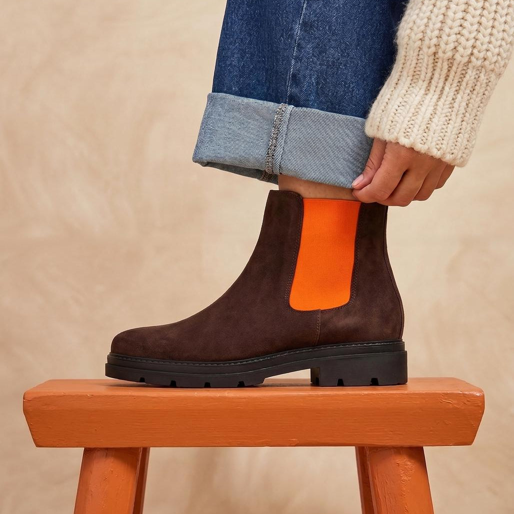 A person wearing a dark brown suede Chelsea boot with a bright orange elastic side panel, paired with blue jeans and a cream knit sweater, sitting on a wooden stool against a textured beige background.