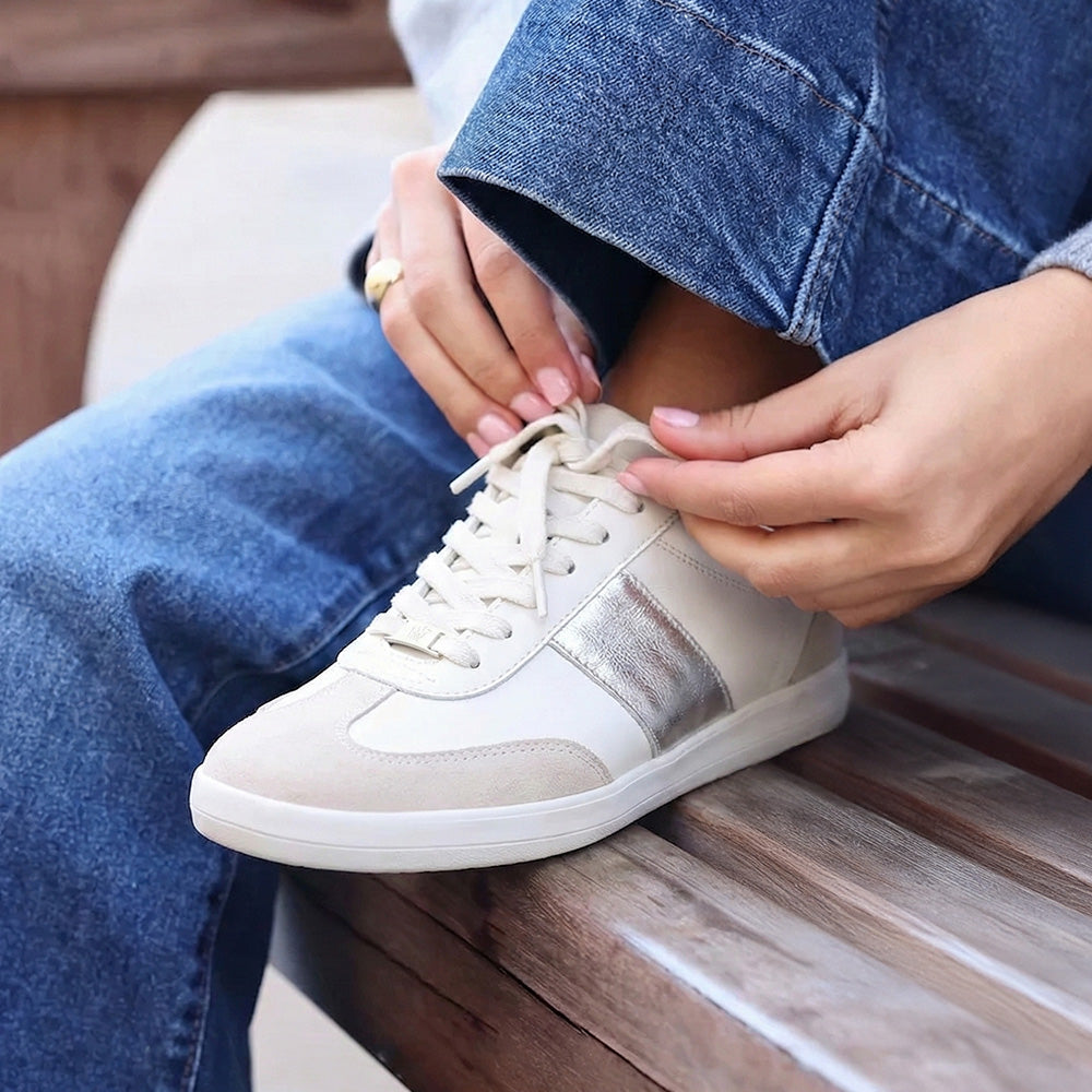 Person tying a white sneaker with a metallic stripe on a wooden bench.