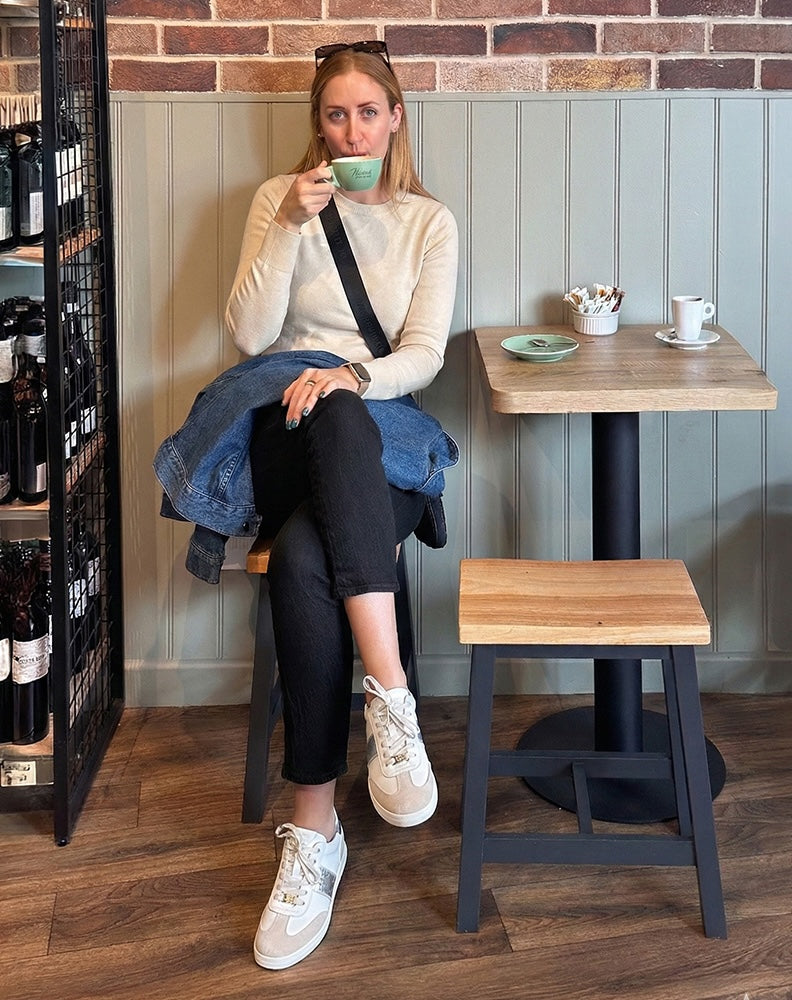 Woman sitting at a cafe table drinking coffee, wearing white leather sneakers with a brick wall and wooden floor in the background.