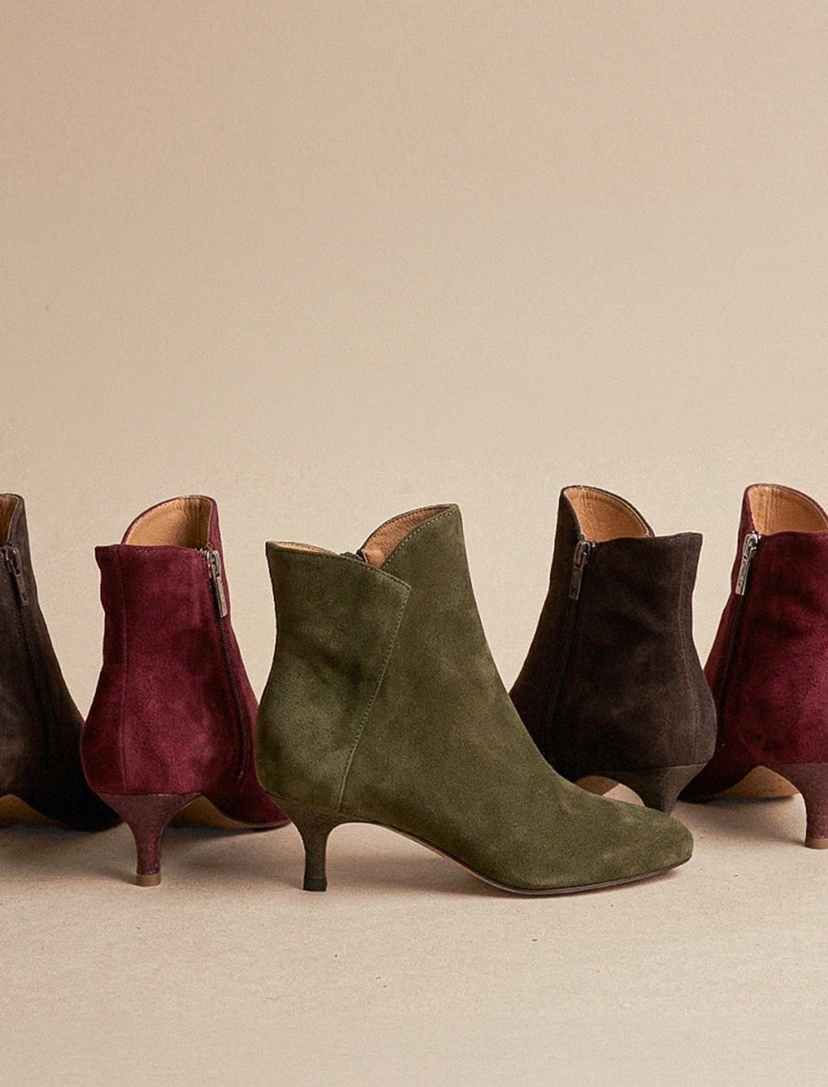 Close-up of two suede ankle boots on a white background, one in navy and one in tan brown, both featuring a side zipper and a large fringed detail on the outer side.
