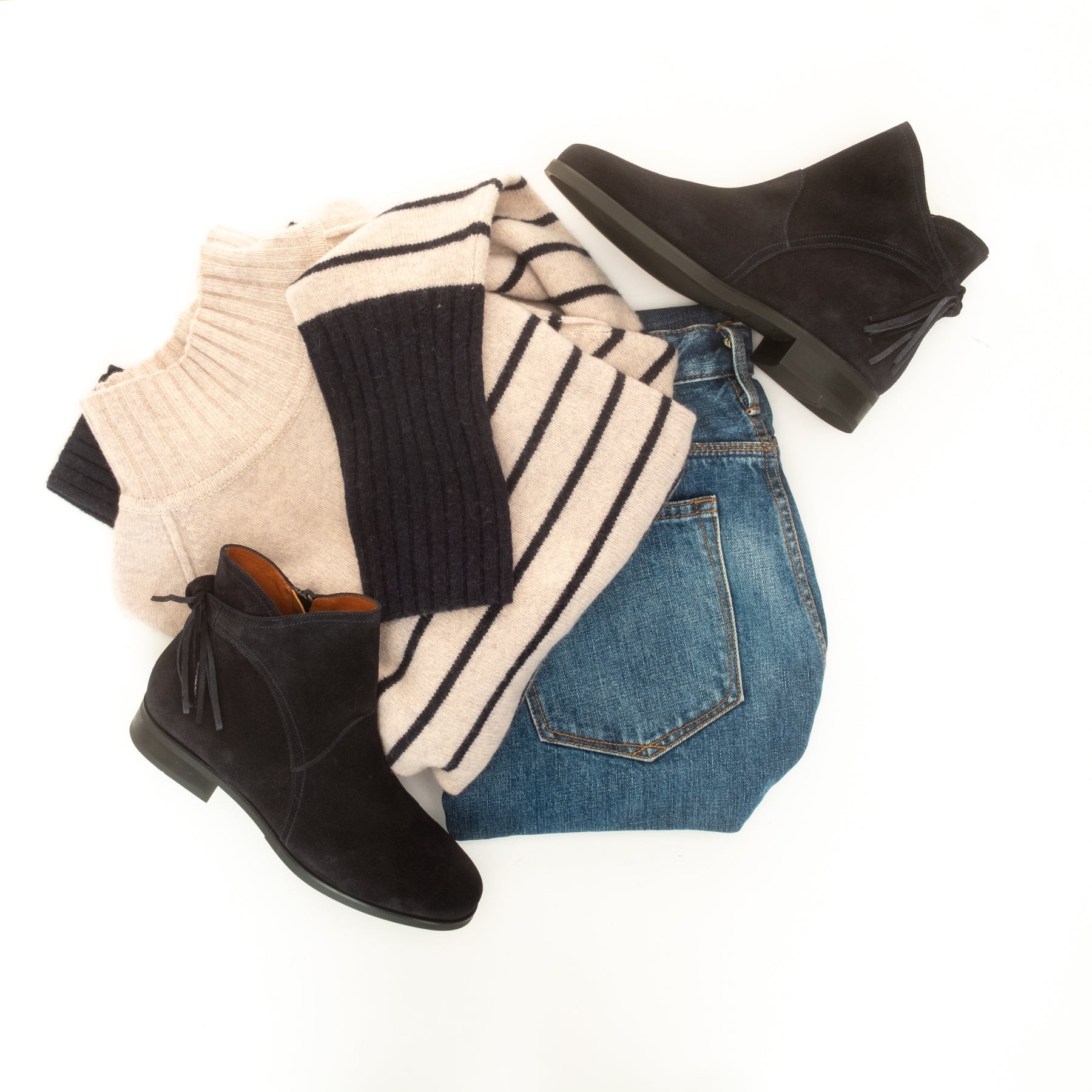 A styled flat lay of women's fall/winter clothing on a white background, including black suede ankle boots, blue jeans, a cream and navy striped knit sweater, and a cream ribbed turtleneck.
