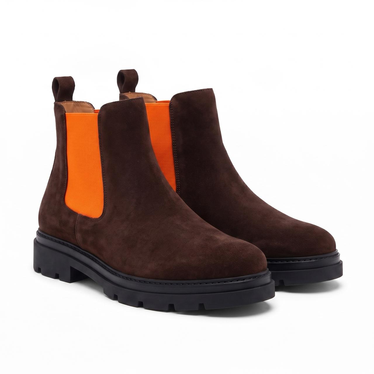 A pair of dark brown suede Chelsea boots with bright orange elastic side panels and chunky black lug soles, shown on a clean white background.
