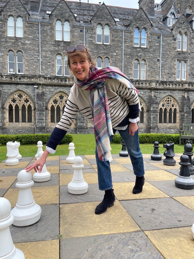 A woman wearing a striped sweater, blue jeans, and a red plaid scarf poses on a stone chessboard in front of a large historic stone building.