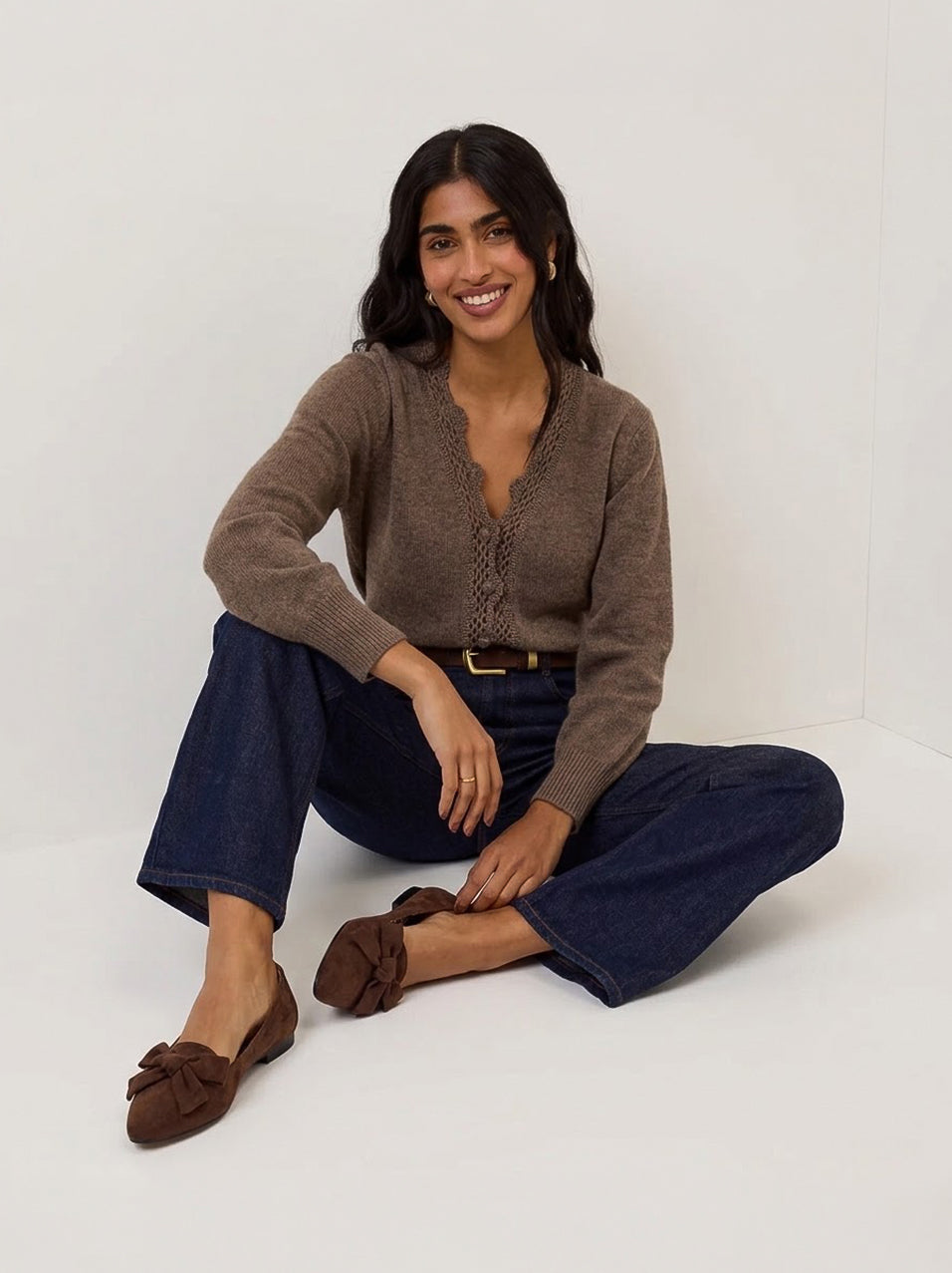 A woman sits cross-legged on a white studio floor wearing a brown knit cardigan, dark blue cropped jeans, and brown suede loafers.