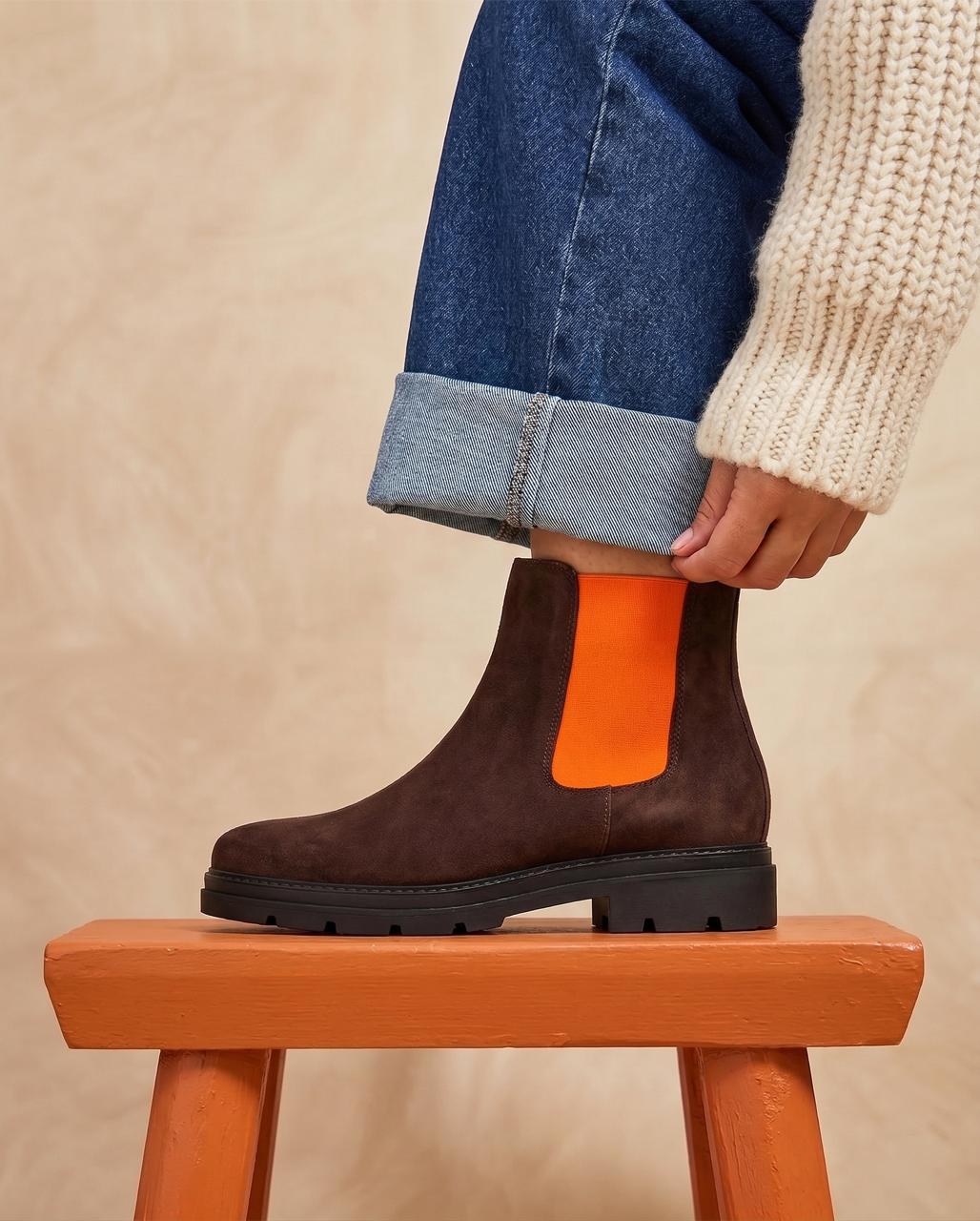 A person wearing a dark brown suede Chelsea boot with a bright orange elastic side panel, paired with blue jeans and a cream knit sweater, sitting on a wooden stool against a textured beige background.