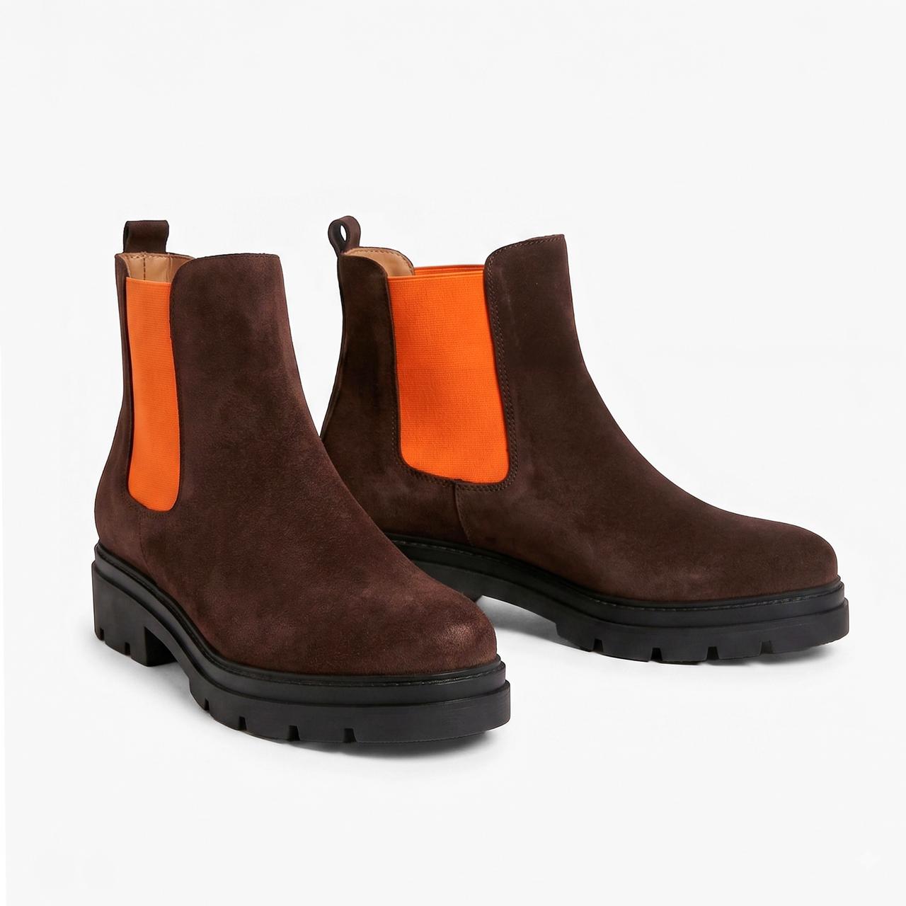 A pair of dark brown suede Chelsea boots with bright orange elastic side panels and chunky black lug soles, shown on a clean white background.