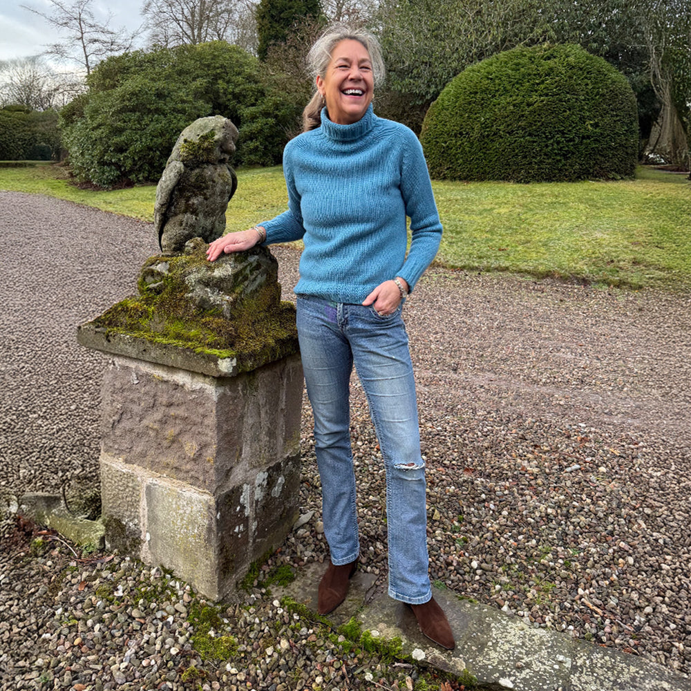Woman in a blue sweater standing next to a stone lion statue in a garden.