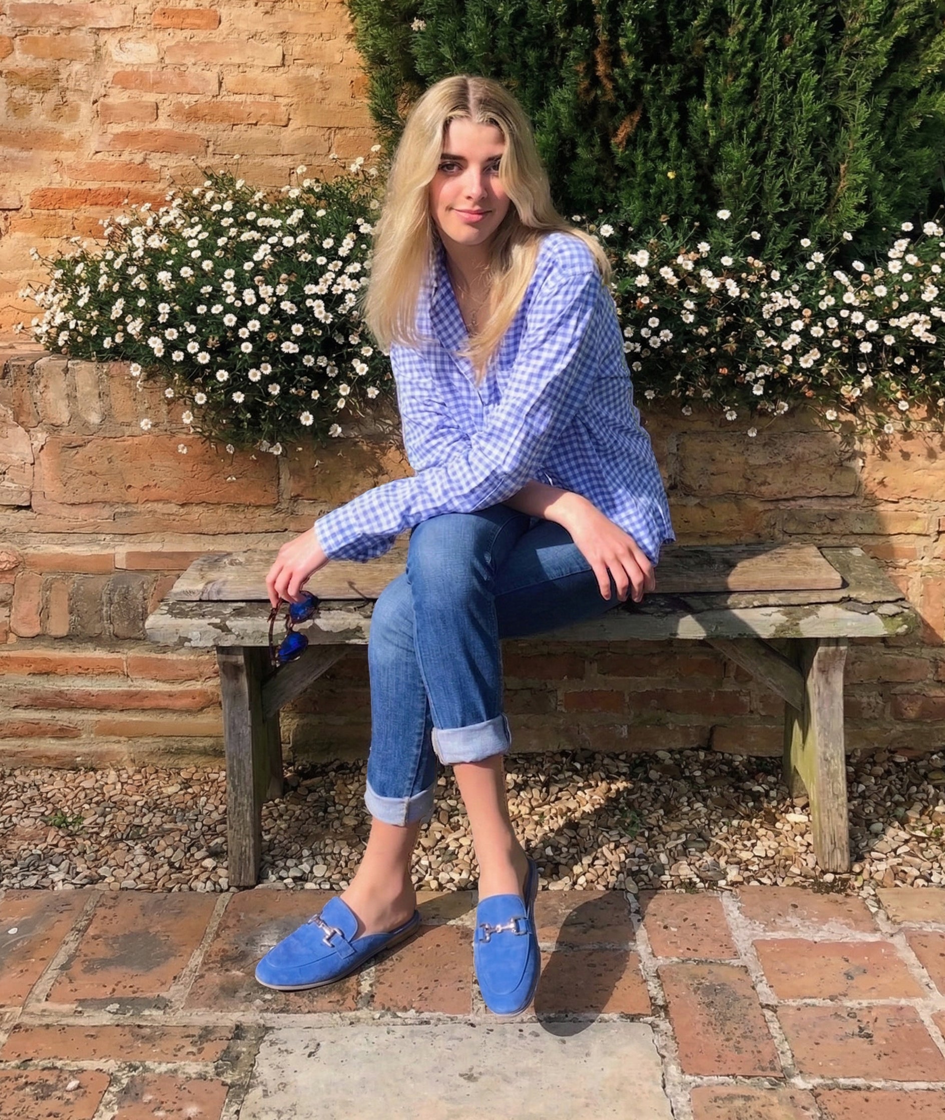 Woman sitting on a wooden bench wearing a blue checkered shirt and blue suede mules, with a brick wall and flowers in the background.