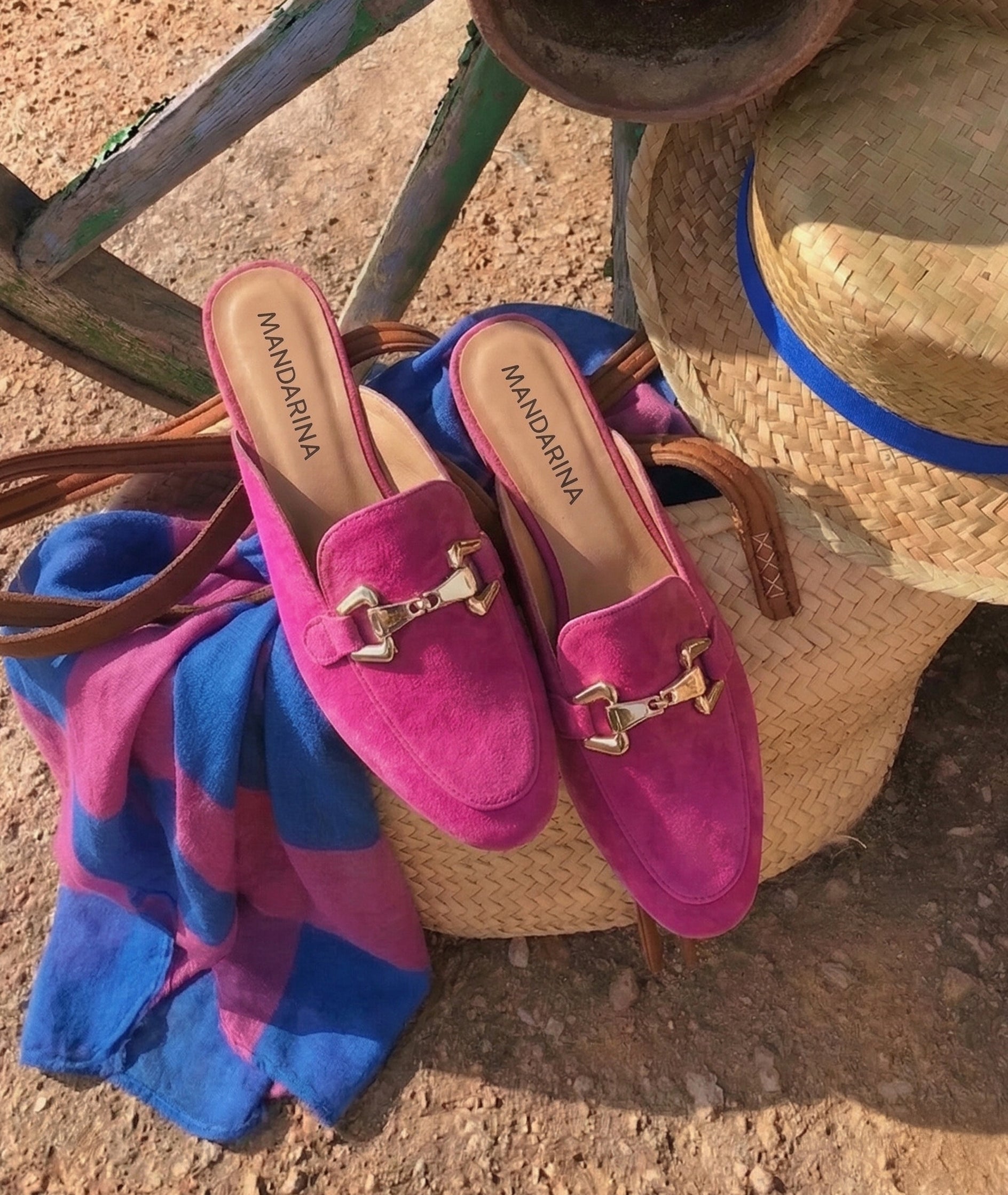 Pink suede shoes with buckles on a blue and pink striped bag, next to a straw hat on a textured surface.