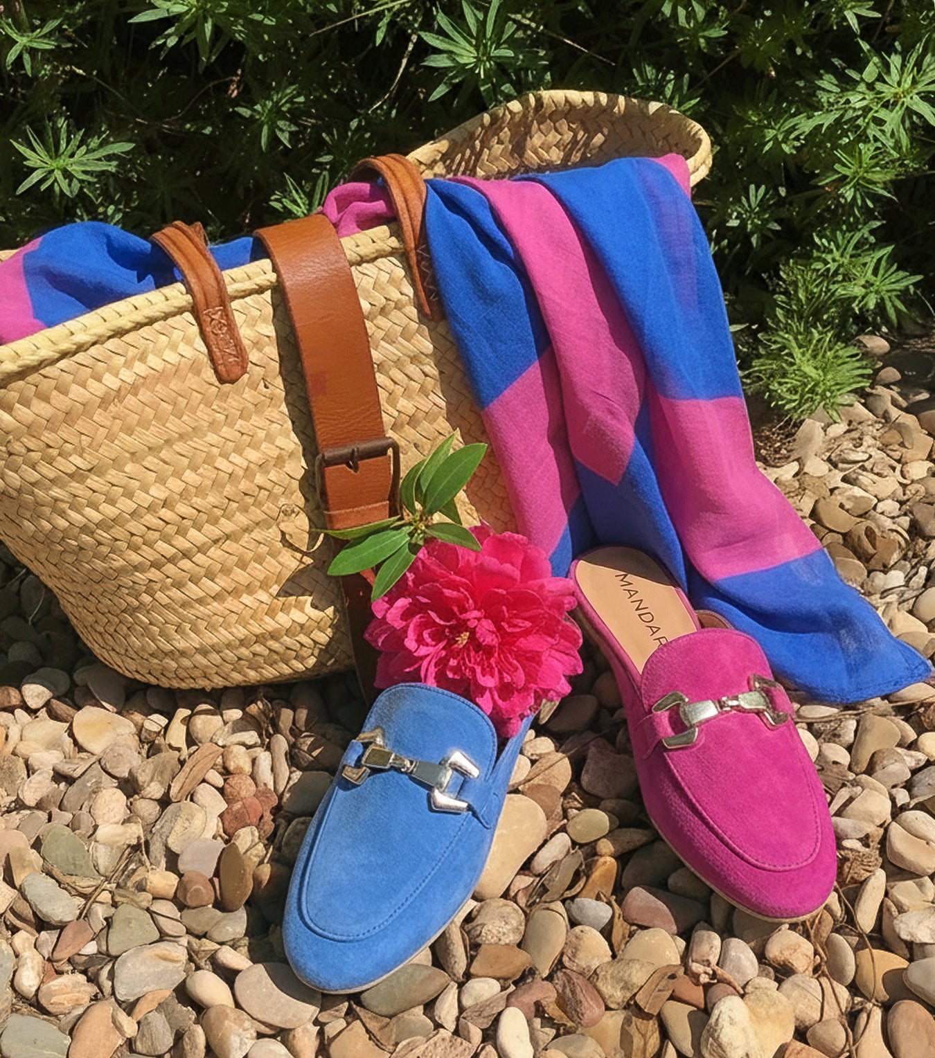 Colorful loafers with a straw bag and pink flower on pebbles